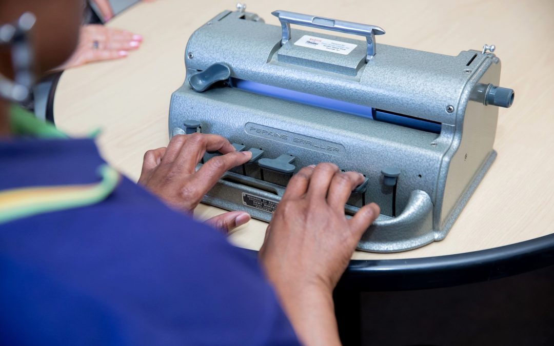 picture taken over the shoulder of a person wearing a purple polo shirt. They are typing on a Brailler.