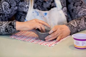 A woman is seated at a table. Only her torso, arms and hands are visible. She is wearing a black and white, paisley patterned long-sleeve blouse. She has a ring on her left ring finger. She is molding a bowl out of grey clay with her hands. The clay sits on a square of fabric, that has a pattern comprised of red, light grey and pink half-circles. An empty purple and white cup is by the woman's left hand.