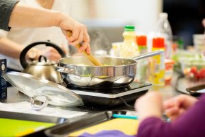 The picture focuses on a low pot, placed on a hot plate. A person's hand is seen holding a wooden spoon, stirring the contents of the pot. The lid of the pot is in the foreground and different cooking items like oils and sprays are in the background.