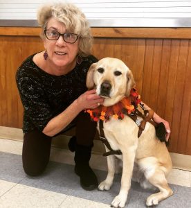 A woman with short, curled blonde hair, is kneeling down on her right knee. She is wearing burgundy tights and a 1/2 sleeve black dress with white and red patterns. She has on purple dangling earrings and black-rimmed glasses. She is smiling, holding on to the harness of her guide dog, who is sitting. The guide dog is a yellow labrador retriever with some white and light brown pigment in her fur. She is wearing a dog harness, along with a festive collar. The collar is made up from orange and red sheer, triangles with red, yellow, brown and orange turkey shapes attached to the ends of them.