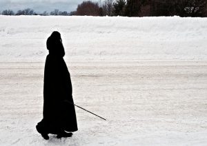 A person, wearing a long dark coat with a hood, is walking on a field of snow, using a white cane to orient.